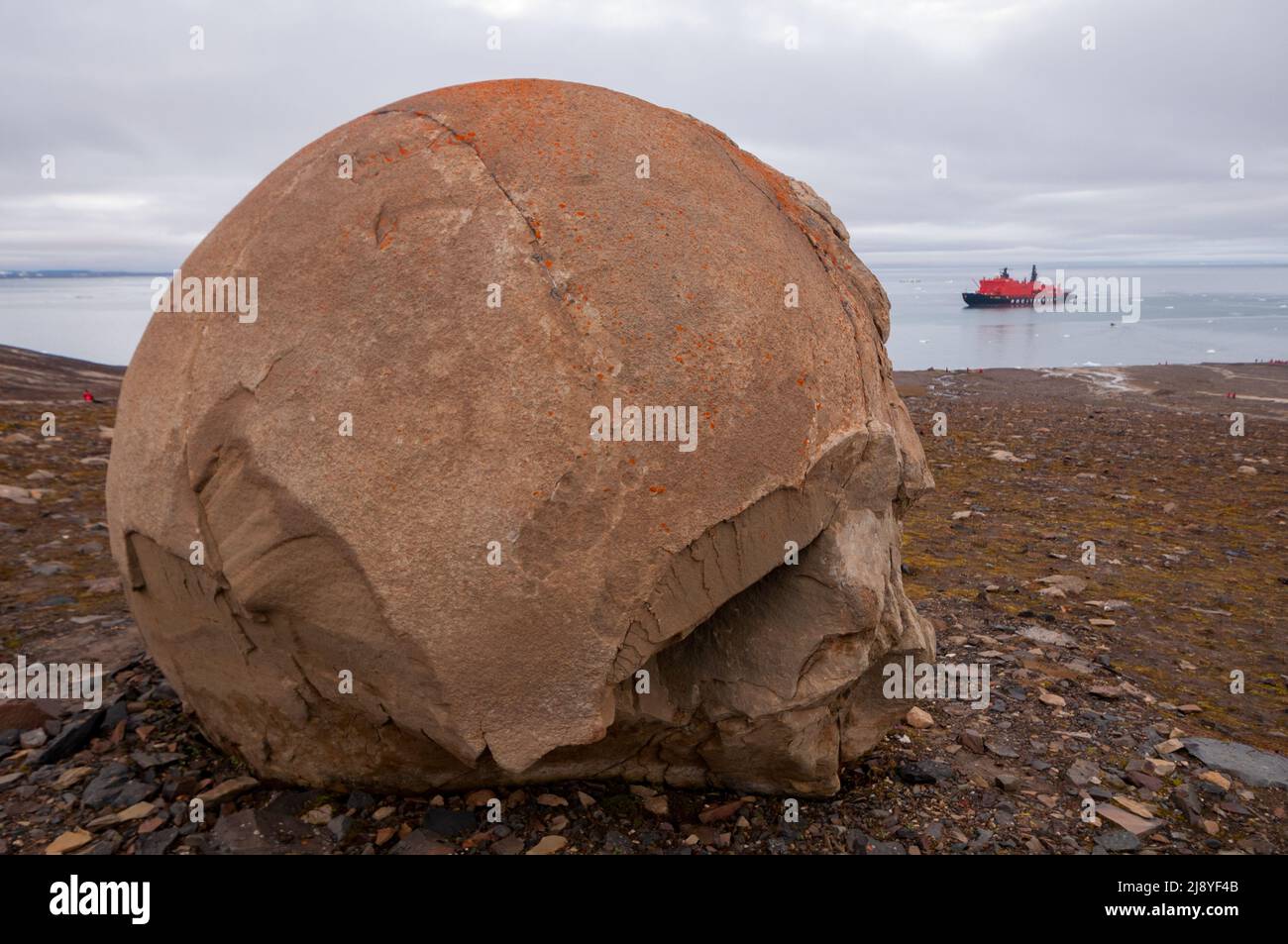 Ein riesiger Felsball unbekannter Herkunft, wahrscheinlich ein Beton, auf Champ Island, Franz Josef Land, Russland, mit Eisbrecher im Hintergrund Stockfoto