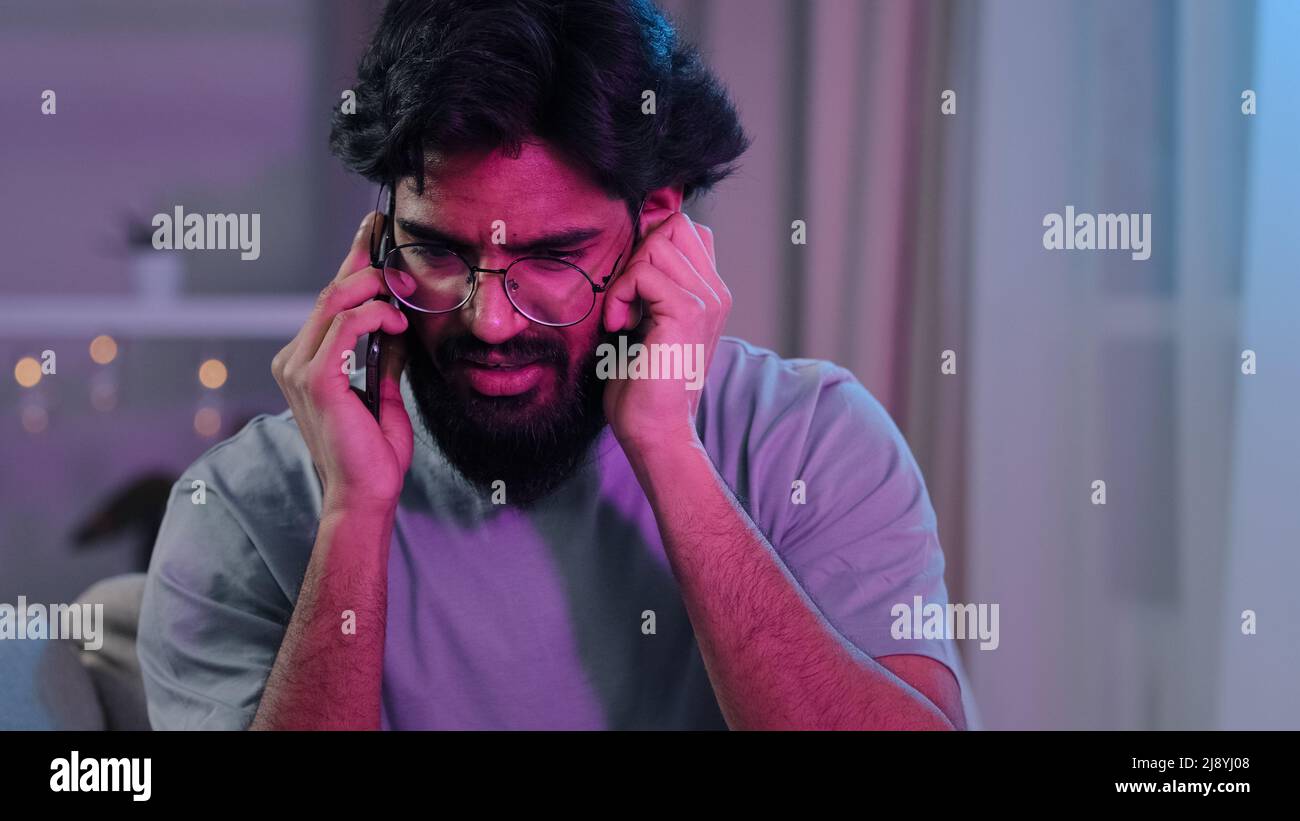 Arabisch-indischer bärtiger Mann mit Brille beim Telefongespräch auf der Hausparty auf dem Sofa sitzen Neonlicht schwer zu hören Unterhaltung laute Musik lärmender Geburtstag Stockfoto