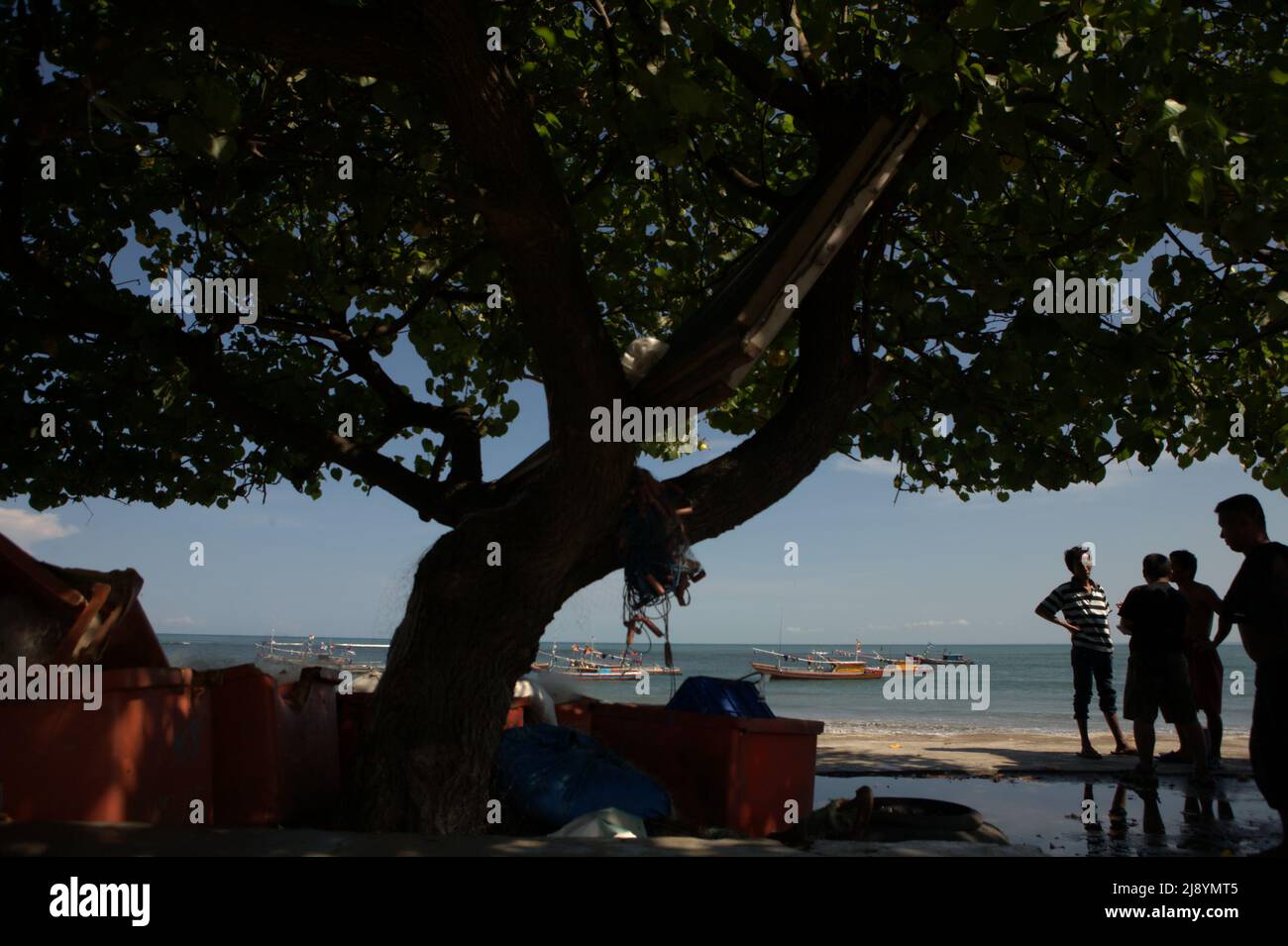 Silhouette von Männern, die sich unter einem Baum im Hintergrund von Fischerbooten auf dem Küstenwasser am Malabero-Strand unterhalten, einem flachen Strand in Bengkulu, Indonesien. Stockfoto