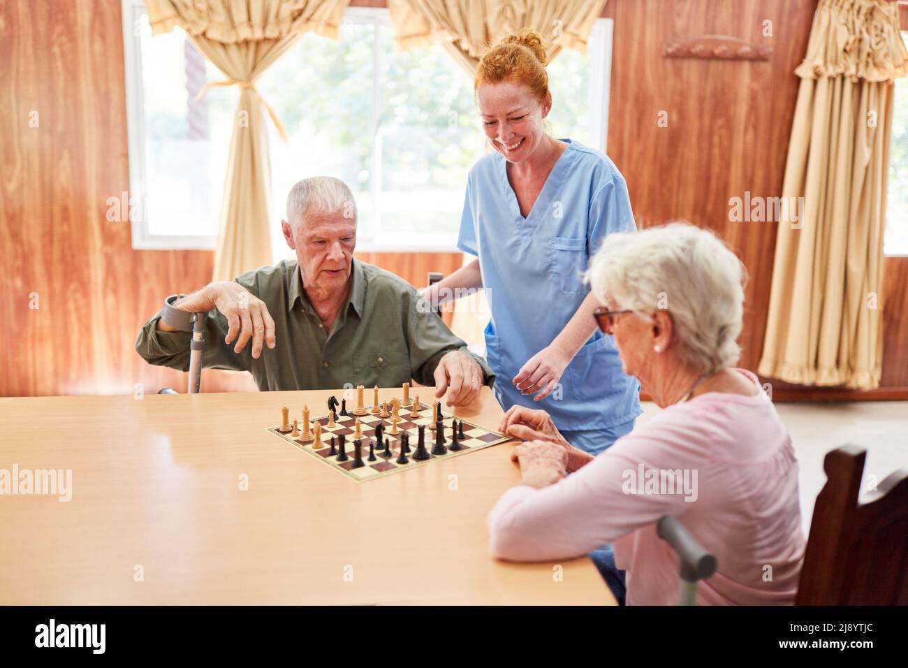 Ein altes Seniorenpaar im Pflegeheim spielt Schach neben einer lächelnden geriatrischen Krankenschwester Stockfoto