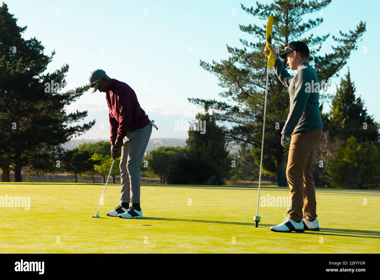 Kaukasischer junger Mann mit Golfflagge, während ein afroamerikanischer Freund Golfball mit Schläger trifft Stockfoto
