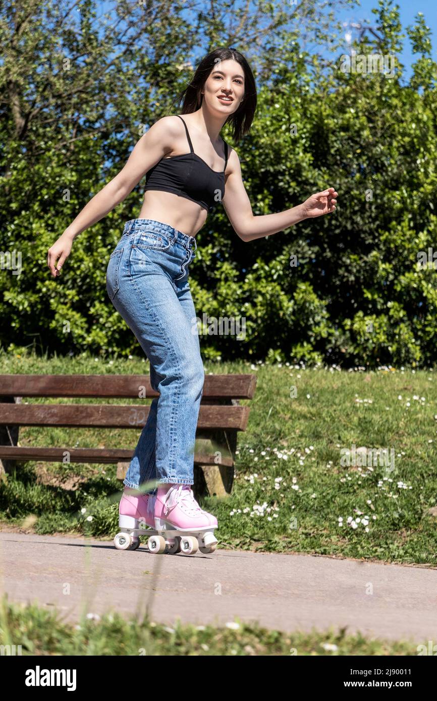 Brunette Frau im Park Skaten trägt Sport-BH, Jeans und rosa Rollschuhe Stockfoto