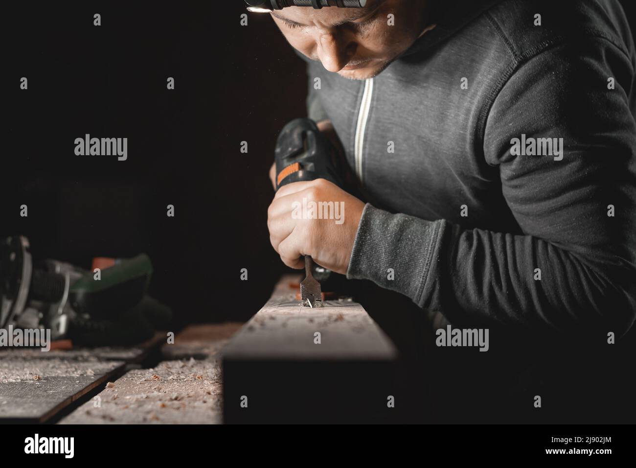 Ein Mann arbeitet mit einer Bohrmaschine in seiner Werkstatt. Zimmermann bohrt mit einem Hand-Elektrowerkzeug in einem dunklen Raum mit gerichtetem Licht Stockfoto