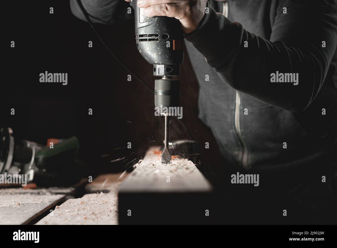 Ein Mann arbeitet mit einer Bohrmaschine in seiner Werkstatt. Zimmermann bohrt mit einem Hand-Elektrowerkzeug in einem dunklen Raum mit gerichtetem Licht Stockfoto