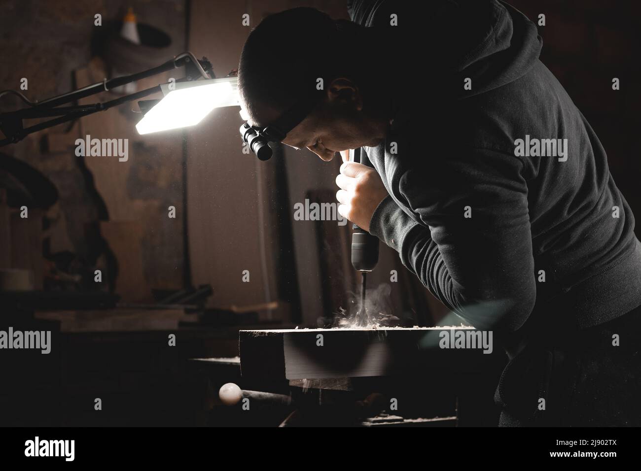 Ein Mann arbeitet mit einer Bohrmaschine in seiner Werkstatt. Zimmermann bohrt mit einem Hand-Elektrowerkzeug in einem dunklen Raum mit gerichtetem Licht Stockfoto