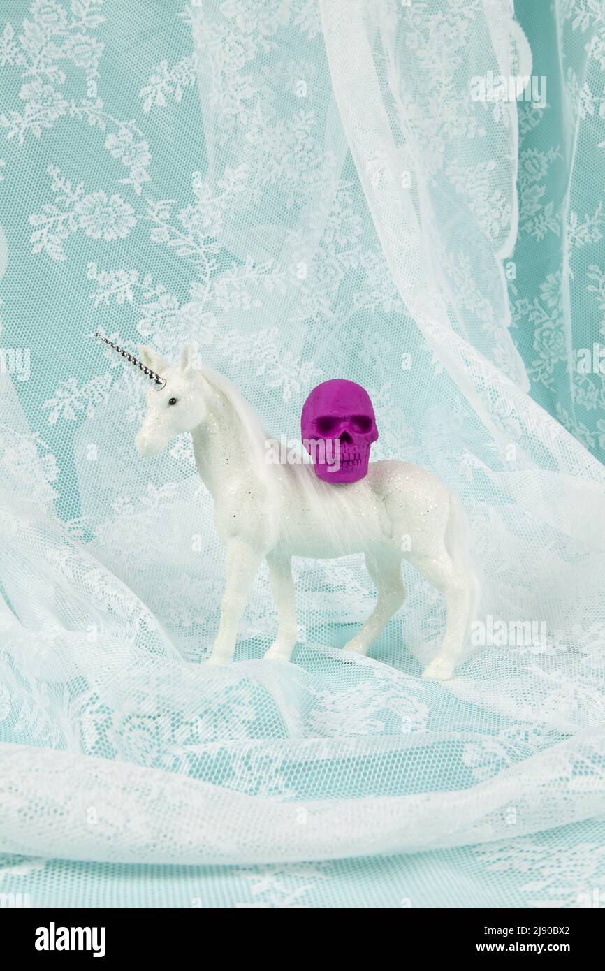 Ein glänzendes weißes Einhorn, das einen Plastikschädel auf dem Rücken trägt. Minimale und kreative Farb-Stillleben-Fotografie Stockfoto