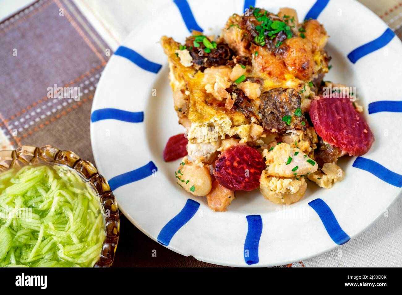 Gebackene Gnocchi mit aufgeschnittener Rote Bete auf gestreiftem Teller und kariertem Tischtuch, Schüssel mit Gurkensalat, Nahaufnahme. Stockfoto