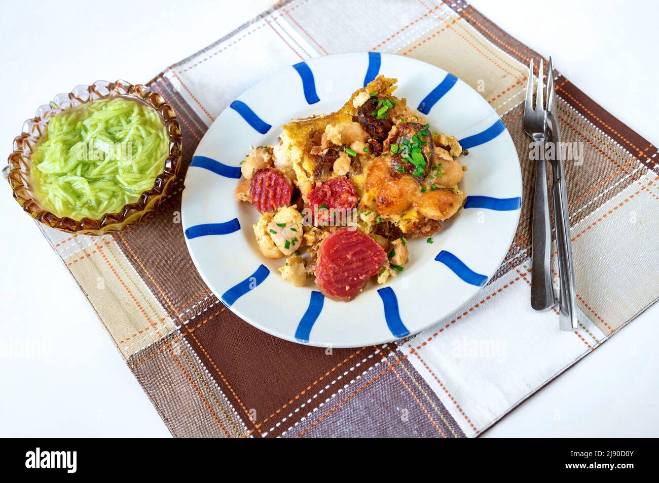 Gebackene Gnocchi mit geschnittener Rote Bete auf gestreiftem Teller und kariertem Tischtuch, Schüssel mit Gurkensalat, Besteck, Serviergröße auf weißem Tisch. Stockfoto