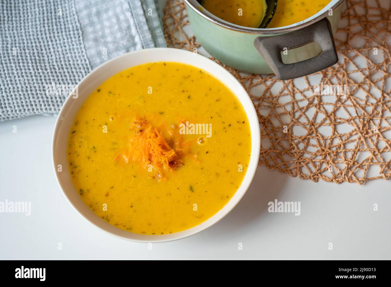 Cremige Karottensuppe auf Teller und in Topf, Handtuch und Bambuskissen auf weißem Tisch. Suppe aus gerösteter Karotte, Gemüsebrühe. Stockfoto