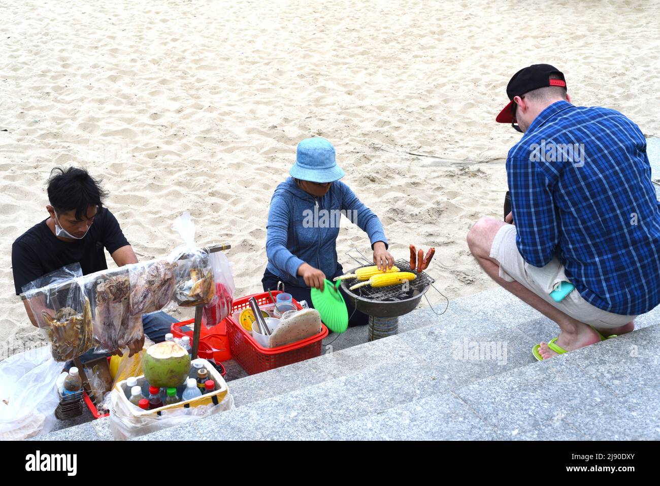 Nha Trang, Vietnam - 17. Mai 2022: Junger Mann bestellt am Strand von Nha Trang von vietnamesen einen gegrillten Mais Stockfoto