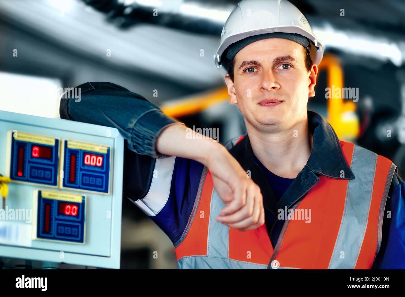 Porträt eines jungen Schwerindustrieingenieurs in Schutzweste und Arbeitskleidung und Helm. Selbstbewusster Mitarbeiter blickt direkt in die Kamera. Moderne technologische Anlage oder Produktionsanlage. Hintergrund Stockfoto