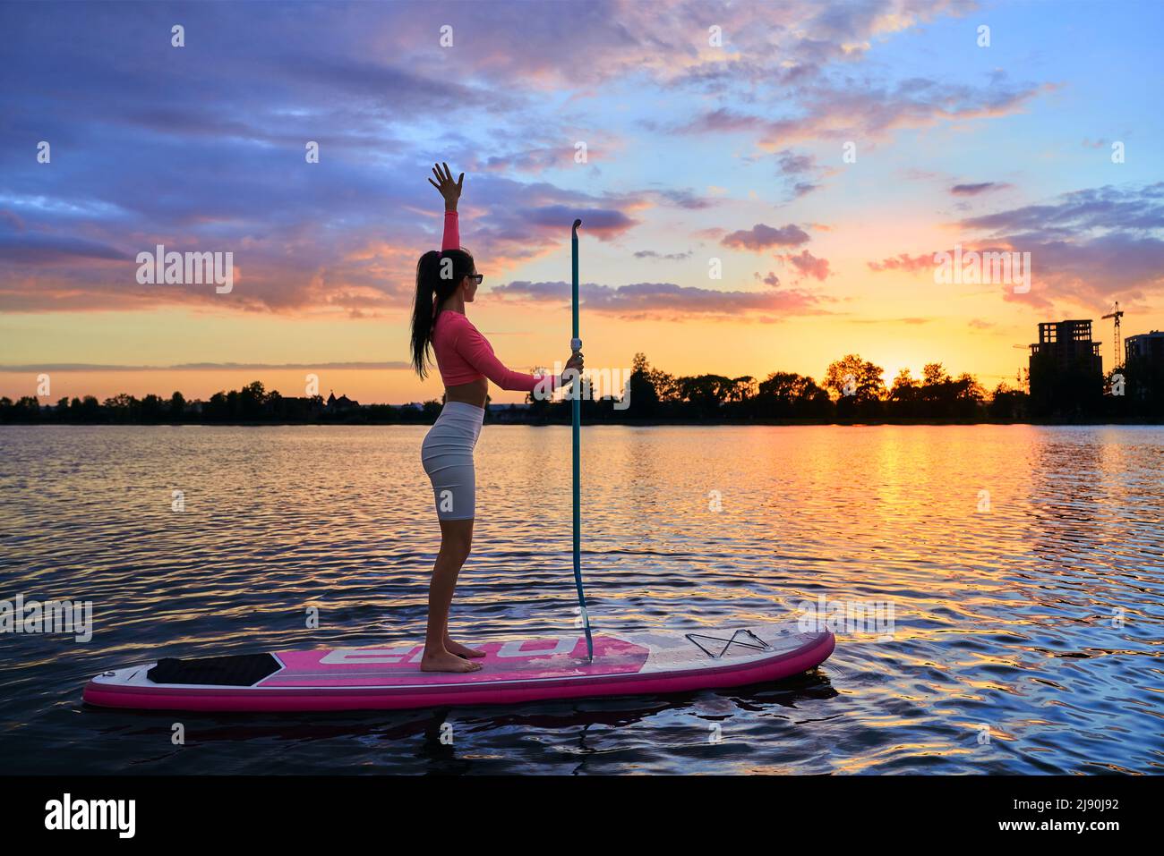 Fitness junge Frau, die mit langem Paddeln in den Händen auf dem Brett steht und den herrlichen Sommeruntergang über dem See genießt. Freizeitaktivitäten im Freien in der Freizeit. Stockfoto