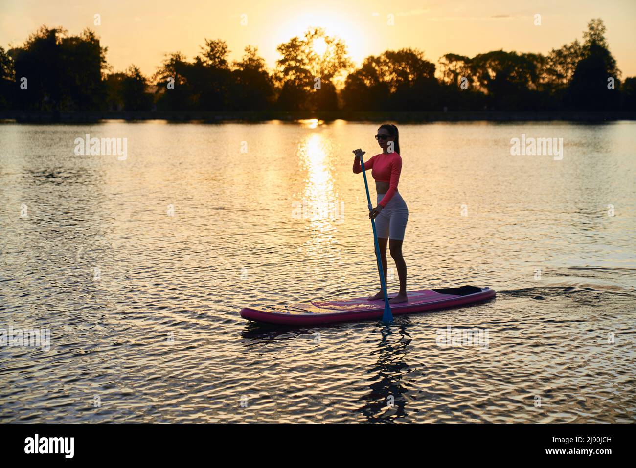 Aktive junge Frau in Sportkleidung und Sonnenbrillen, die abends Zeit zum Üben im Supboarding verbringt. Konzept von Menschen, Freizeit und Wassersport. Stockfoto