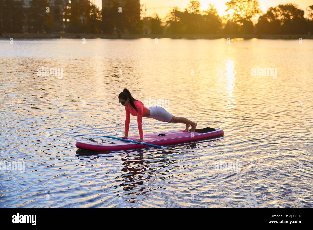 Sportliche kaukasische Frau, die Plank-Übungen macht, während sie auf dem Paddle-Board auf dem Stadtsee schwimmt. Schöner Sommeruntergang im Hintergrund. Gesunde und aktive Lebensweise. Stockfoto