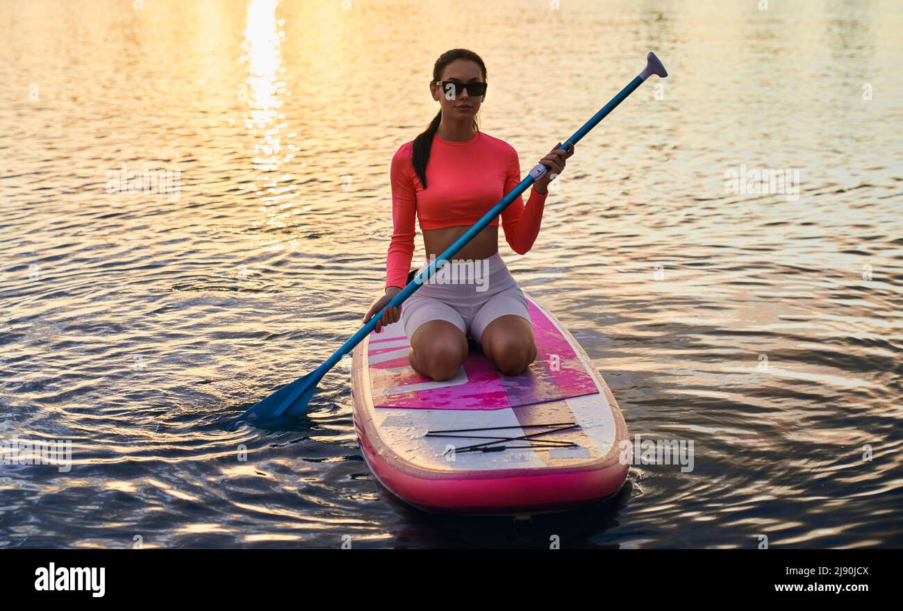 Junge kaukasische Frau, die auf dem Brett sitzt und langes Paddel hält. Attraktive Brünette in Sonnenbrillen und Sportswear mit abendlichen Workout auf City Lake. Stockfoto