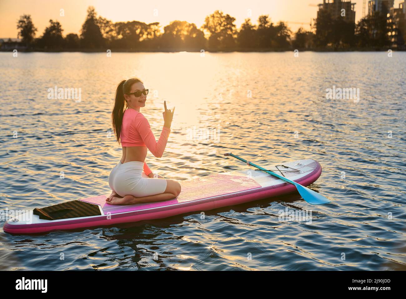 Positive kaukasische Frau sitzt auf dem Brett und zeigt Horngesten auf der Kamera. Glücklich aktive Brünette in Sportswear und Sonnenbrillen genießen Lieblings-Hobby auf City Lake. Stockfoto