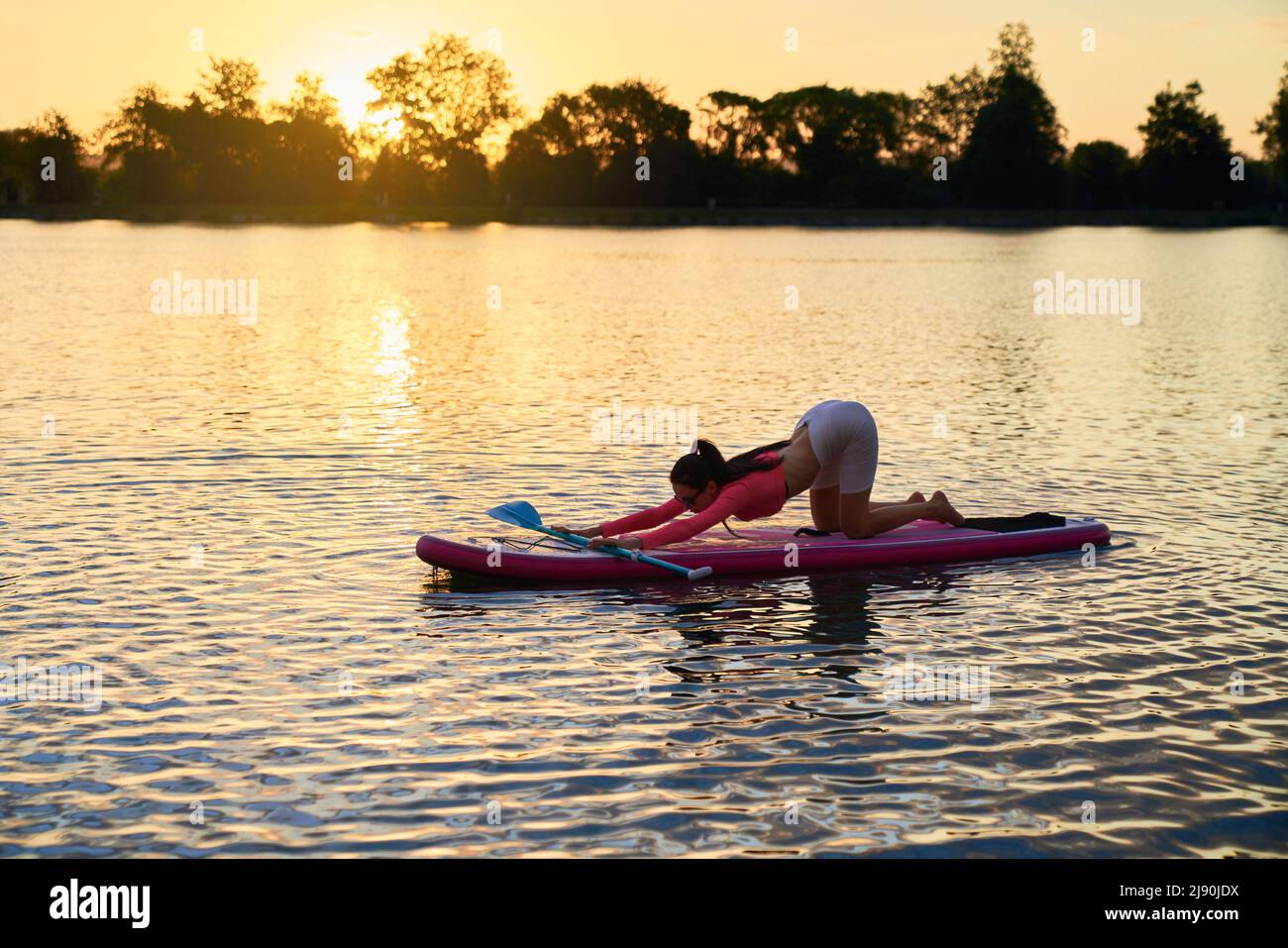 Sportliche junge Frau, die während der Wasseraktivität flexible Übungen macht. Aktive kaukasische Brünette praktiziert Yoga auf dem Brett während des Sonnenuntergangs im Sommer. Stockfoto