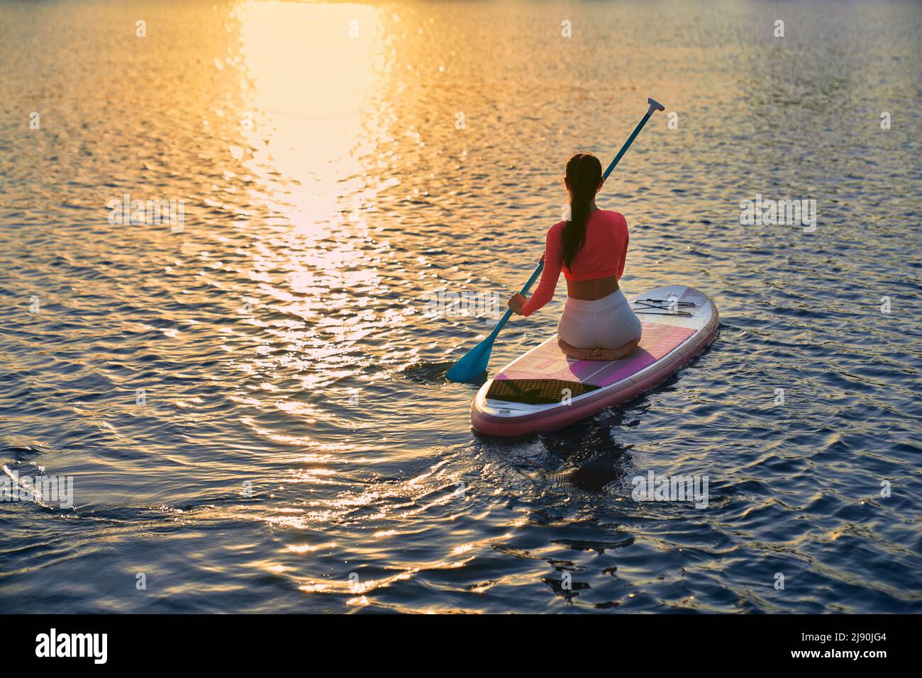 Junge Sportlerin, die am Abend am Stadtsee zum Training im Supboarding unterwegs ist. Schöner Sonnenuntergang im Hintergrund. Reflexion auf dem Wasser. Stockfoto