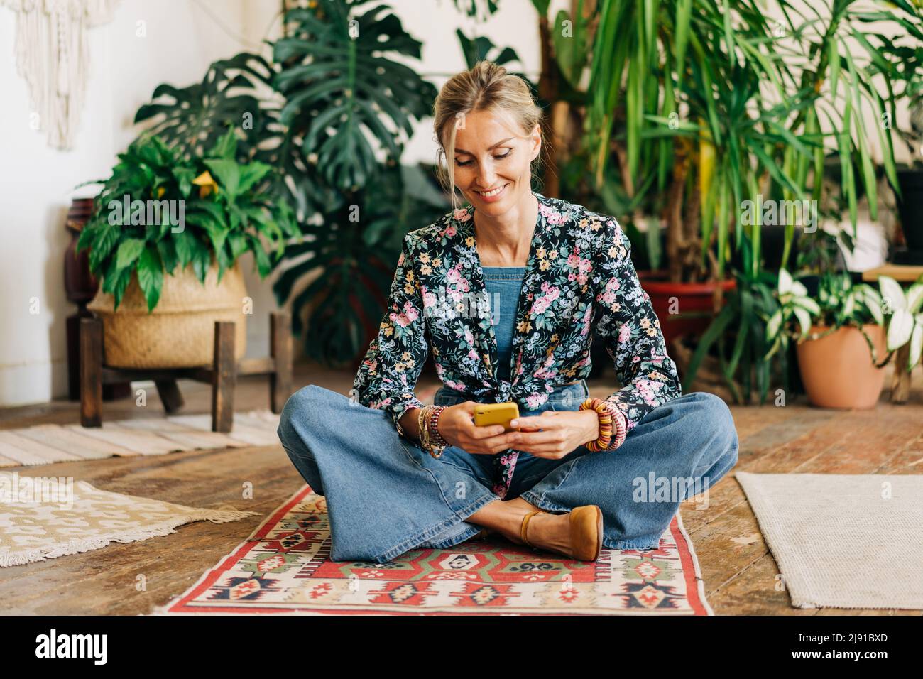 Fröhliche lächelnde Frau, die mit gekreuzten Beinen auf dem Boden sitzt und einen Nachrichtenvorschub auf ihrem Telefon liest. Stockfoto