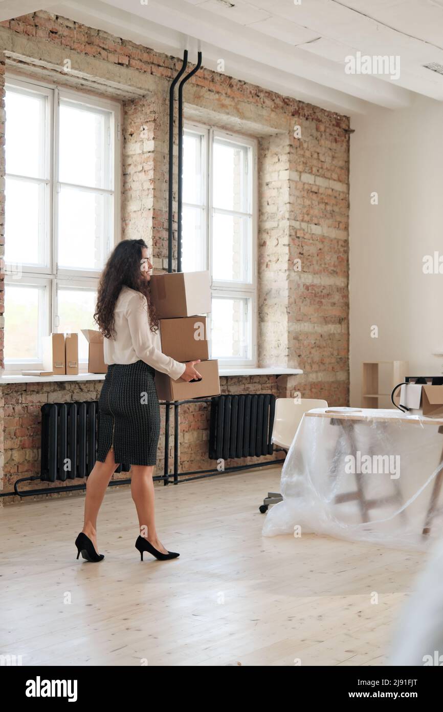 Lächelnde erfolgreiche Geschäftsfrau mit langen lockigen Haaren, die ihre eigenen Sachen in Schachteln verpackt an einen neuen Arbeitsplatz trägt, während sie das Büro oder die Firma wechselt Stockfoto