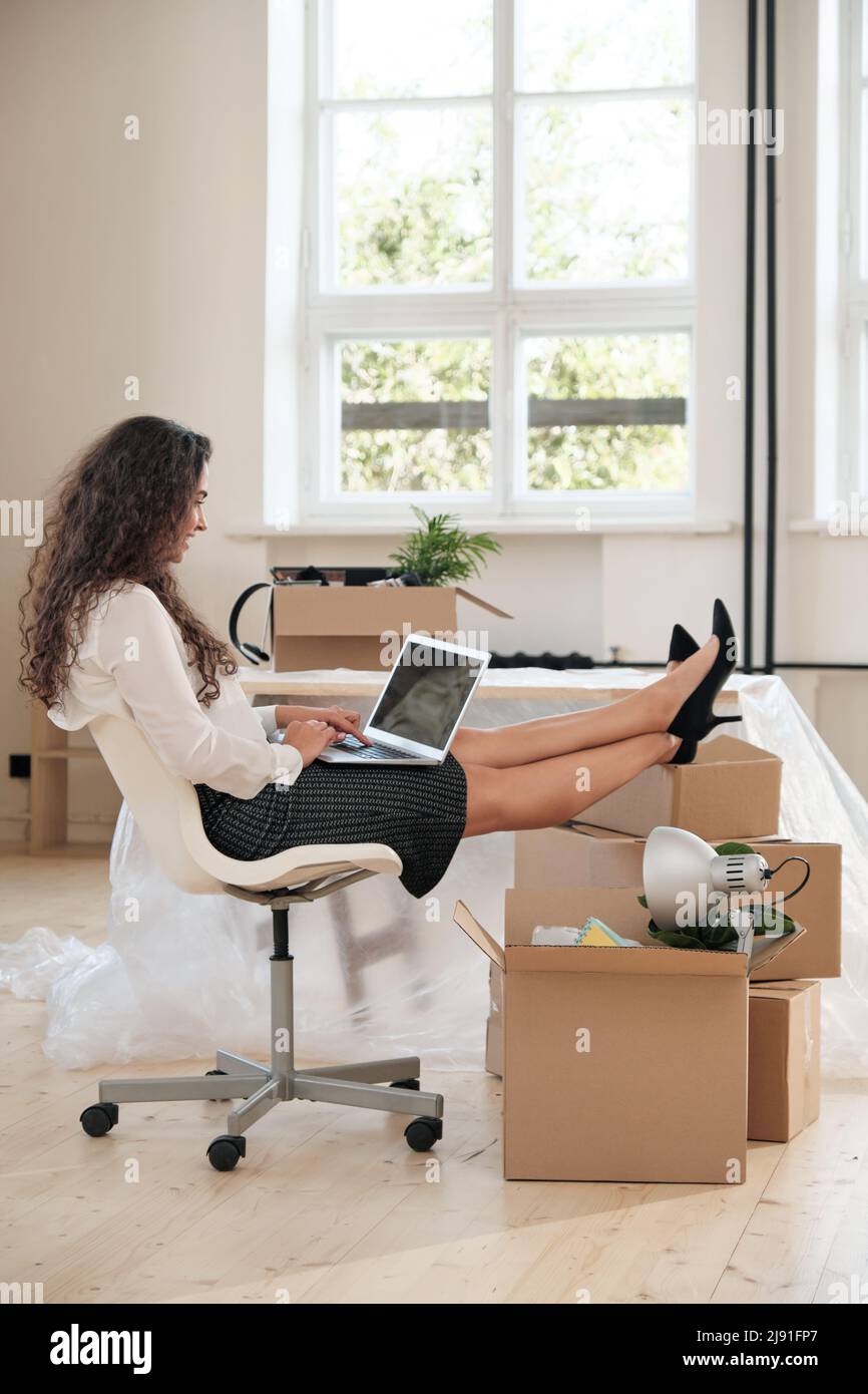 Positive junge Geschäftsfrau im Rock sitzt mit den Füßen auf einem Stapel von Umzugskartons und arbeitet mit einem Laptop in einem neuen Büro Stockfoto