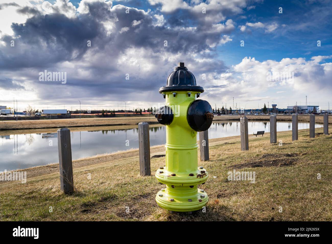 Ein gelber Hydrant, der mit einer Infrastruktur verbunden ist, die neben einem Sturmhalteteich in einem Industriepark in Airdrie, Alberta, Kanada, unter einem Windstampfen steht Stockfoto
