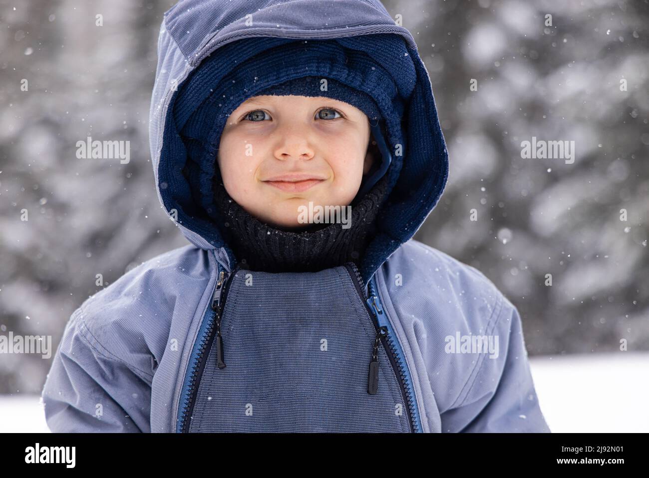 Nahaufnahme eines niedlichen, gesunden 2-jährigen Jungen, warm eingewickelt und im Winter auf dem Land stehend mit verschneiten Bäumen im Hintergrund. Stockfoto