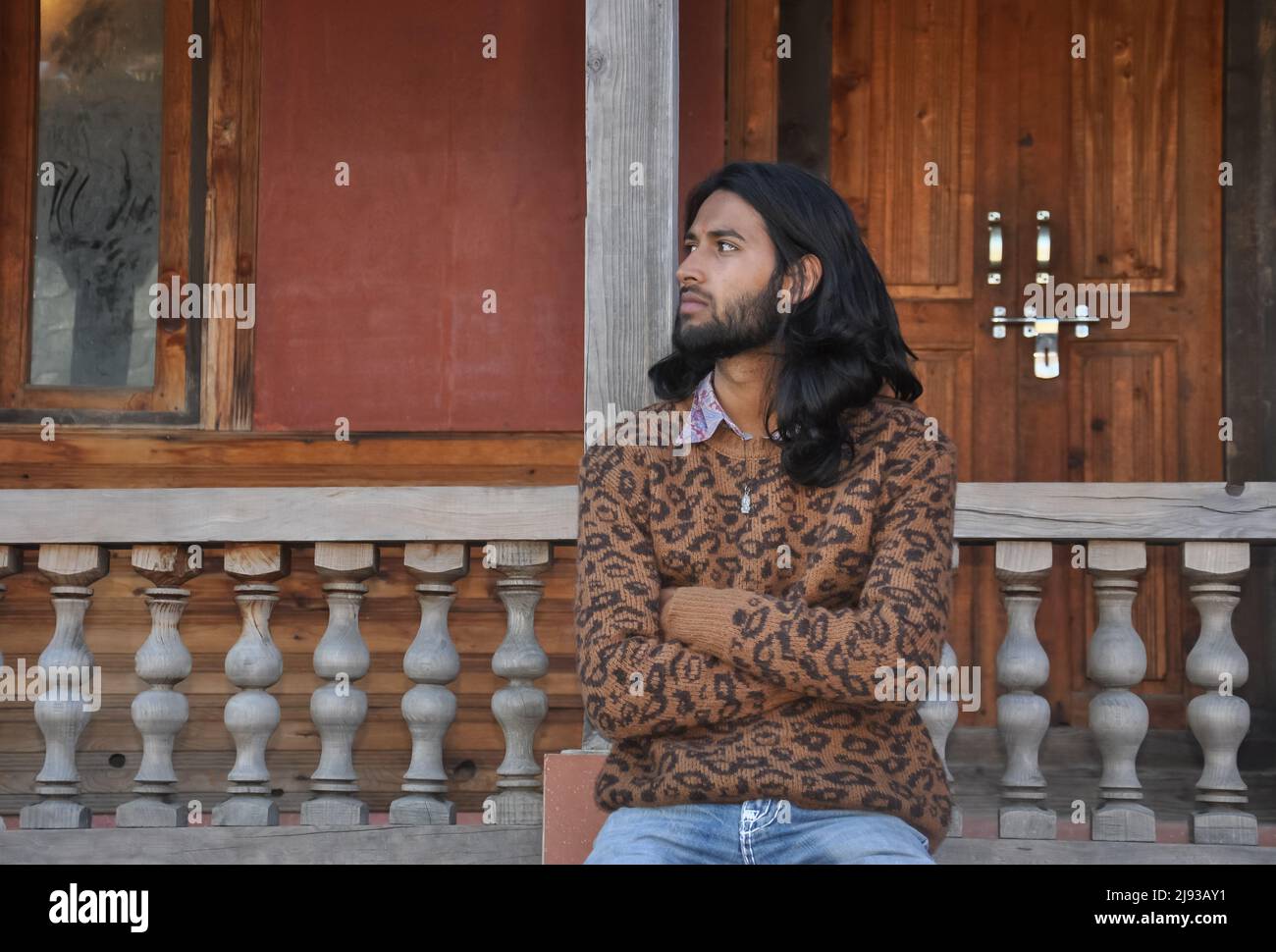 Vorderansicht ein gut aussehender langhaariger junger Kerl, der seitwärts schaut, während er in einem alten Tempel sitzt, der mit gekreuzten Armen posiert Stockfoto