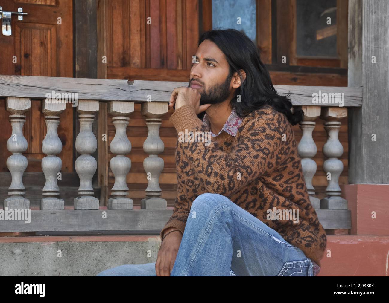 Seitenansicht eines attraktiven langhaarigen indischen jungen Mannes, der zur Seite blickt und mit der Hand auf dem Kinn auf der Tempeltreppe sitzt Stockfoto