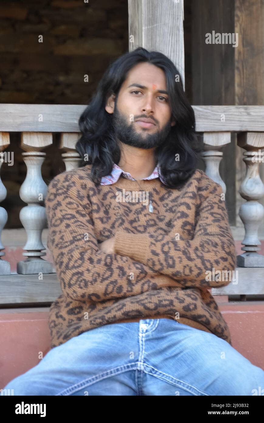 Selektiver Fokus auf einen gut aussehenden jungen indischen Mann mit langen Haaren, der mit gekreuzten Armen posiert und auf die Kamera blickt, während er auf der Tempeltreppe sitzt Stockfoto