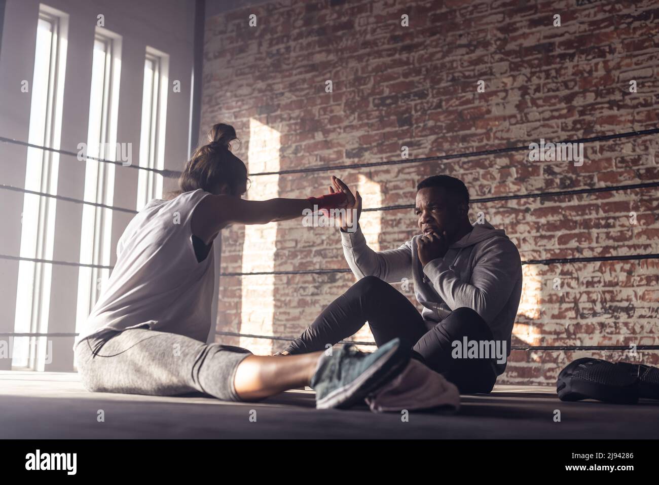 Kaukasische Boxerin, die im Boxring sitzt und mit der Hand des afroamerikanischen männlichen Trainers stanzt Stockfoto