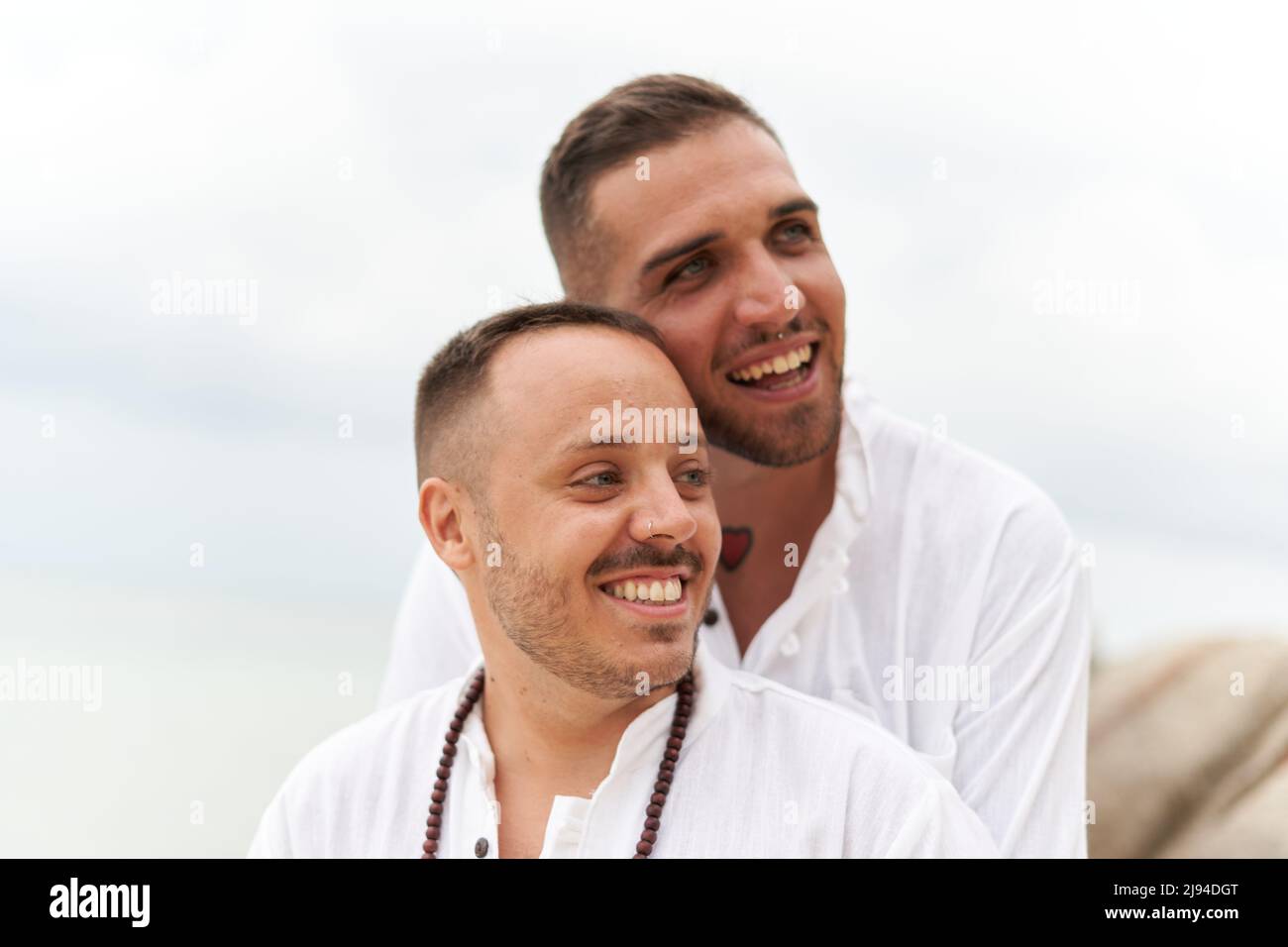 Das homosexuelle Paar lächelt, als es zur Seite schaut und sich am Strand umarmt Stockfoto