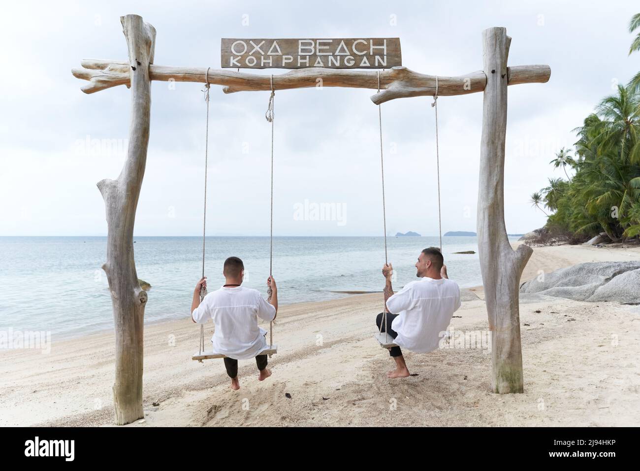 Rücken eines homosexuellen Paares, das auf einer Schaukel an einem tropischen Strand schaukelt Stockfoto