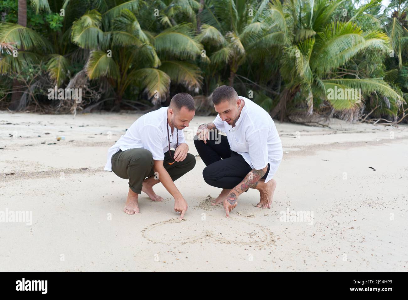 Homosexuelles männliches Paar, das ein Herz im Sand an einem tropischen Strand zieht Stockfoto