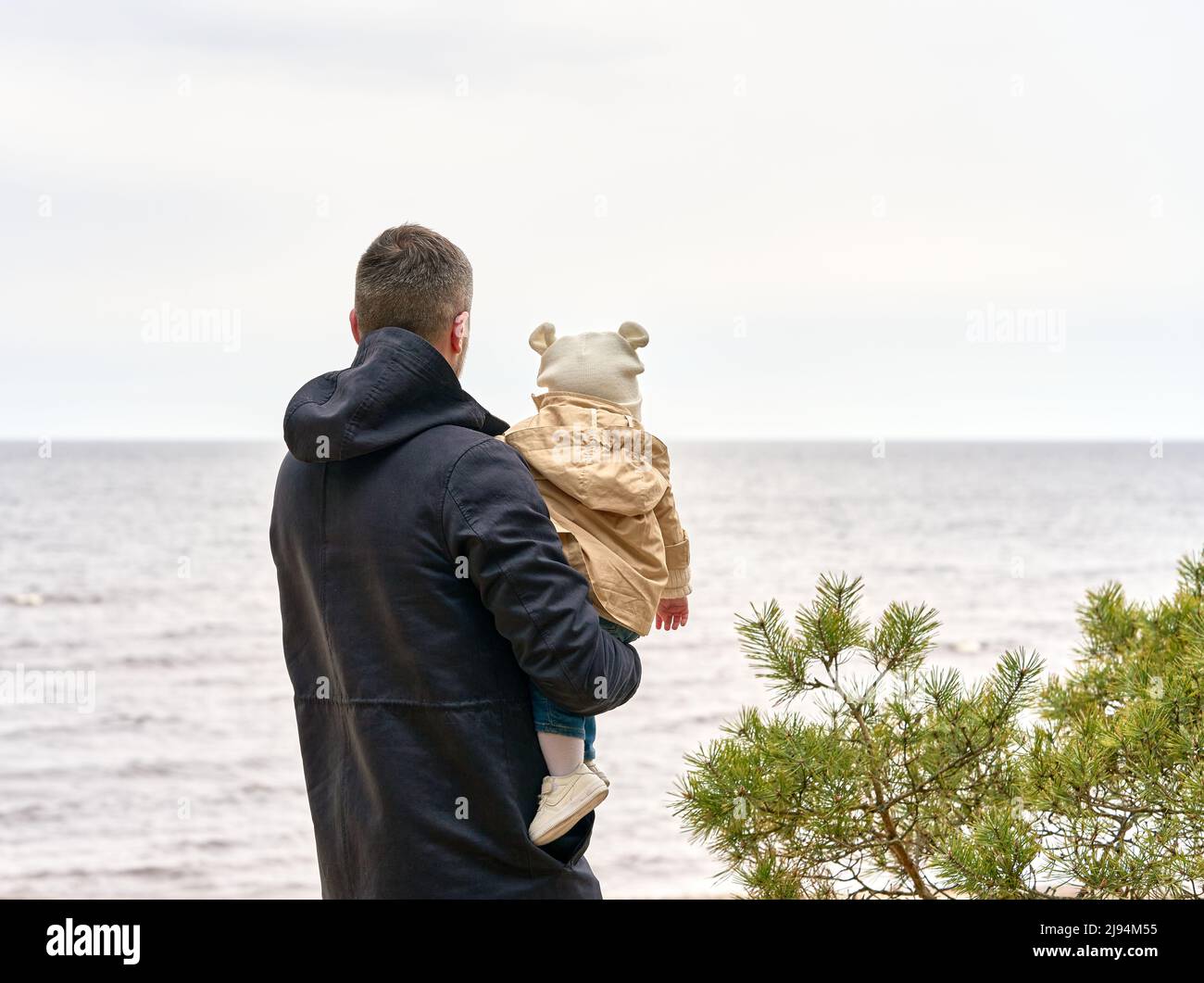 Vater hält Baby auf den Händen. Sie blicken auf den Horizont, der am Ufer des Meeres steht. Kiefer auf der rechten Seite. Stockfoto