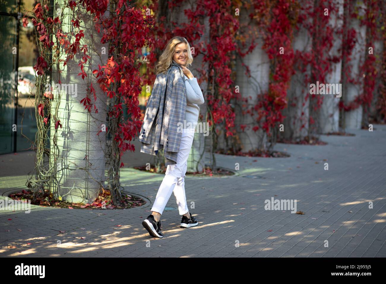 Attraktive blonde 35-40 Jahre alt schaut sich um zu Fuß durch die Herbststadt. Sie ist von Sonnenstrahlen erleuchtet und freut sich über den schönen Tag Stockfoto