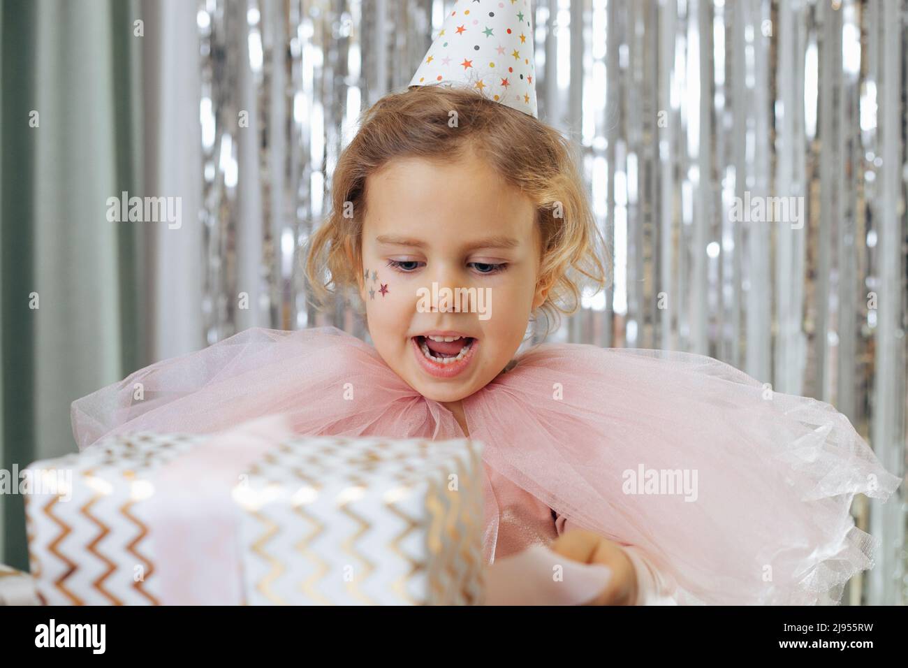 Geburtstag feiern. Fröhlich festlich gekleidet 4-5 - Jahr - altes Mädchen mit Sternen auf ihren Wangen in Party-Hut hält Geschenkbox in ihren Händen. Geschenke für Stockfoto