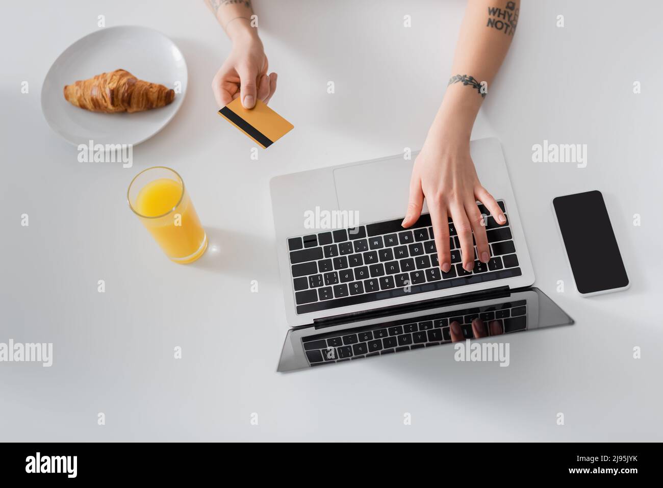 Draufsicht auf eine zugeschnittene Frau mit der Kreditkarte, die auf dem Laptop neben Orangensaft, Croissant und Mobiltelefon tippt Stockfoto