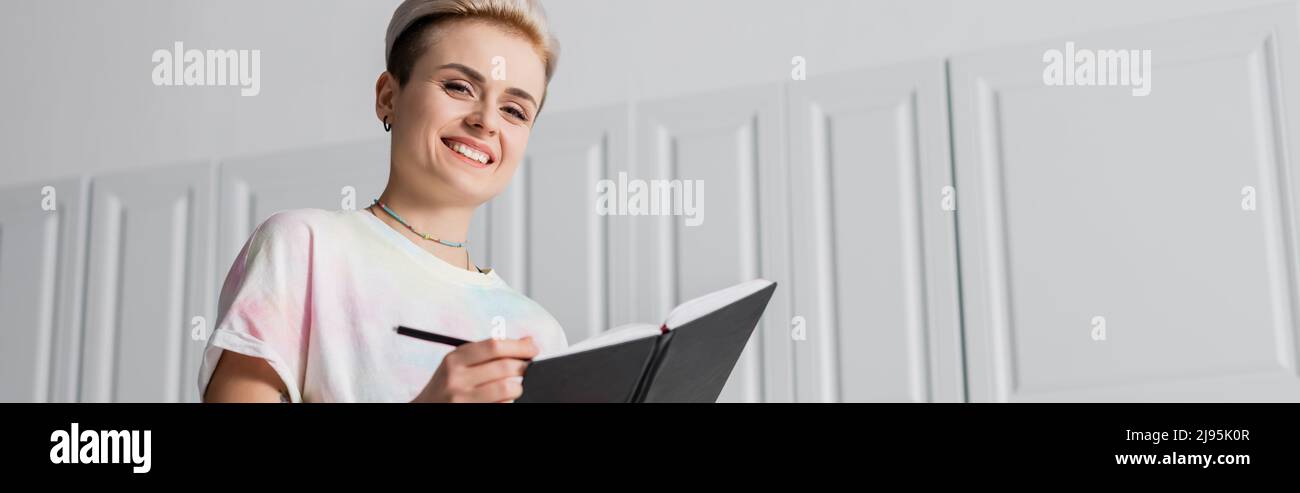 Low-Angle-Ansicht der glücklichen Frau mit trendigen Frisur Schreiben in Notebook, Banner Stockfoto