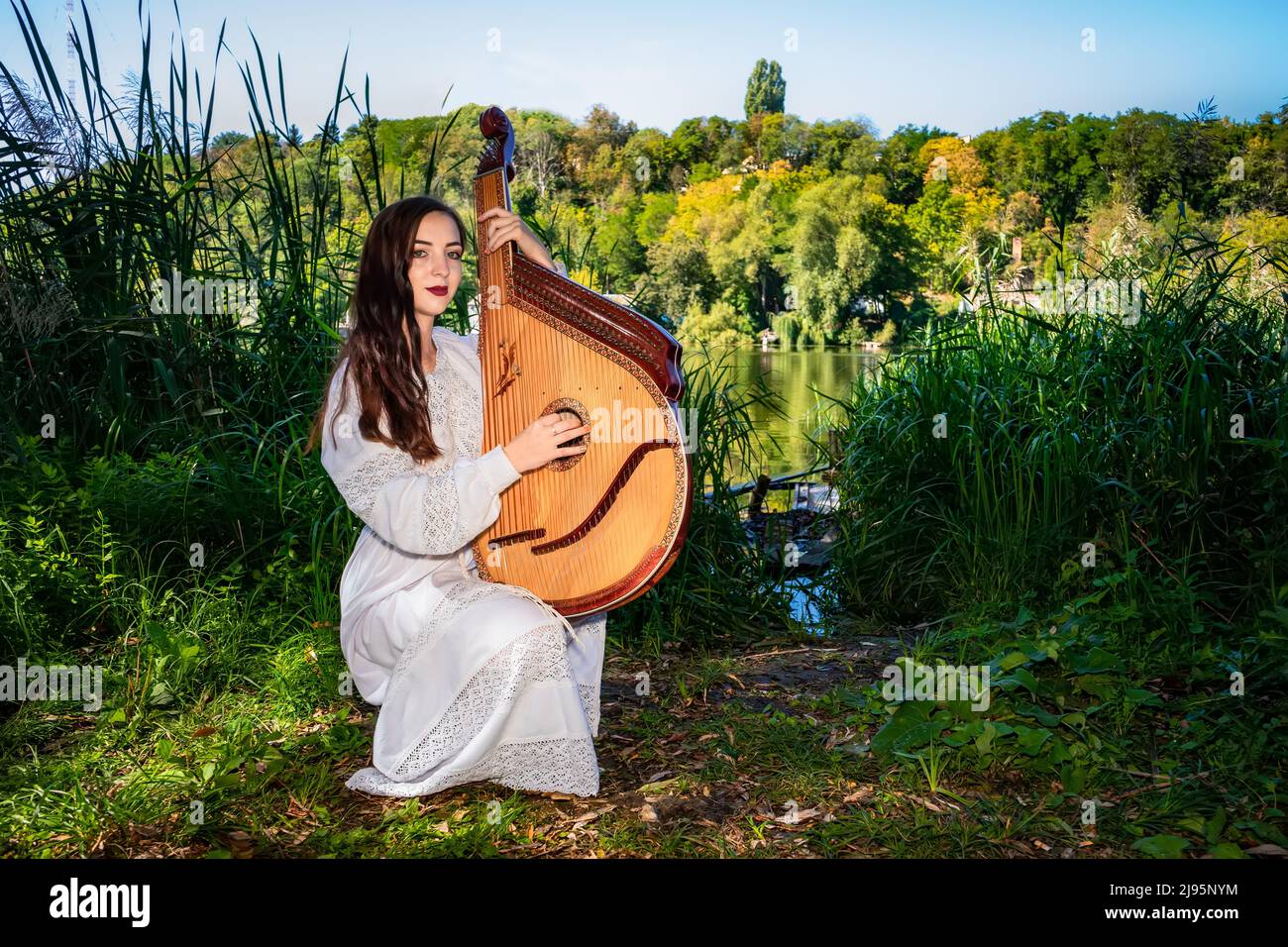 Die junge ukrainische Frau in Nationaltracht spielt an einem sonnigen Sommertag eine Bandura am Fluss. Stockfoto