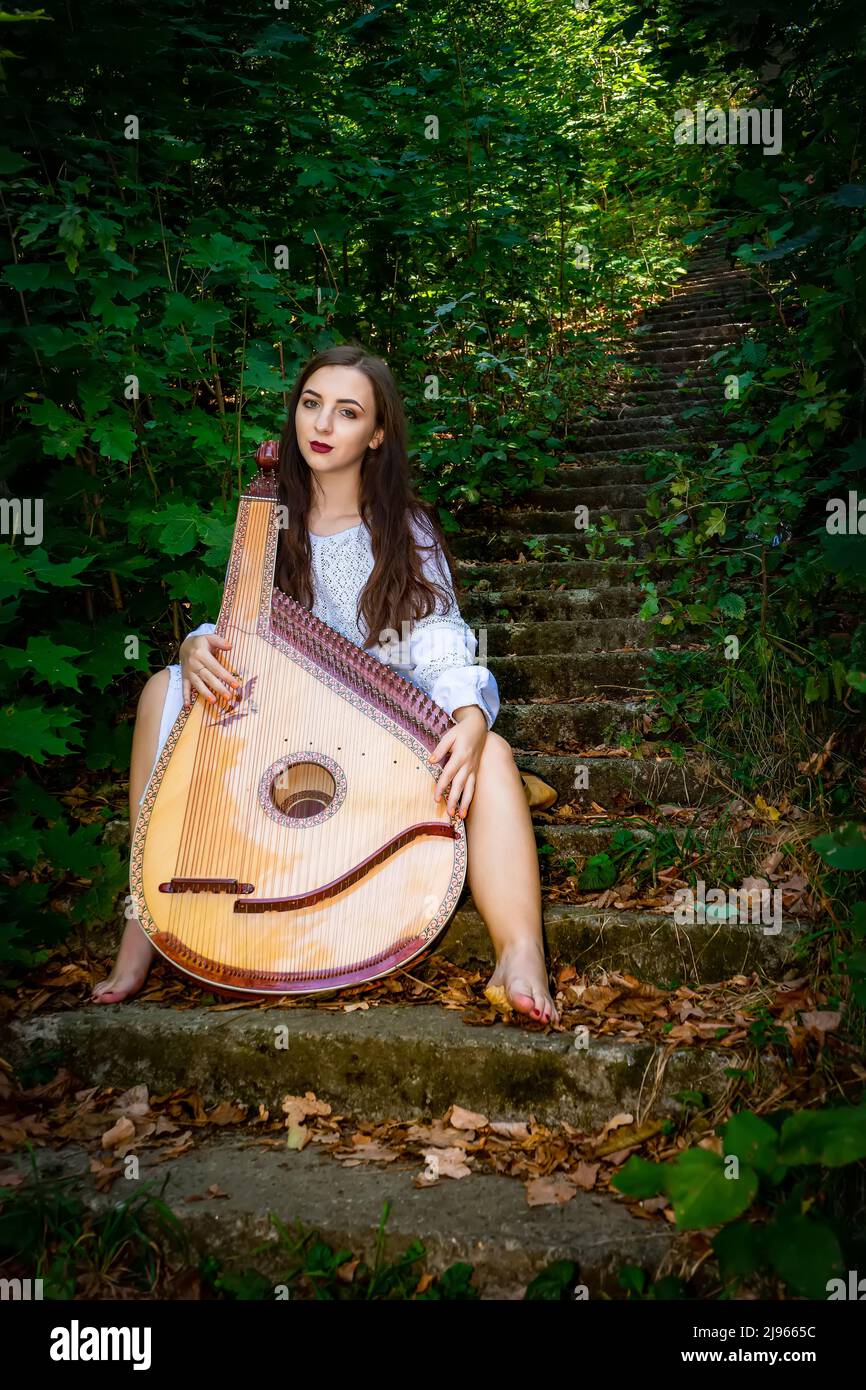 Eine ukrainische Frau sitzt auf der Treppe in einem alten Park mit einem ukrainischen Bandura-Musikinstrument. Stockfoto