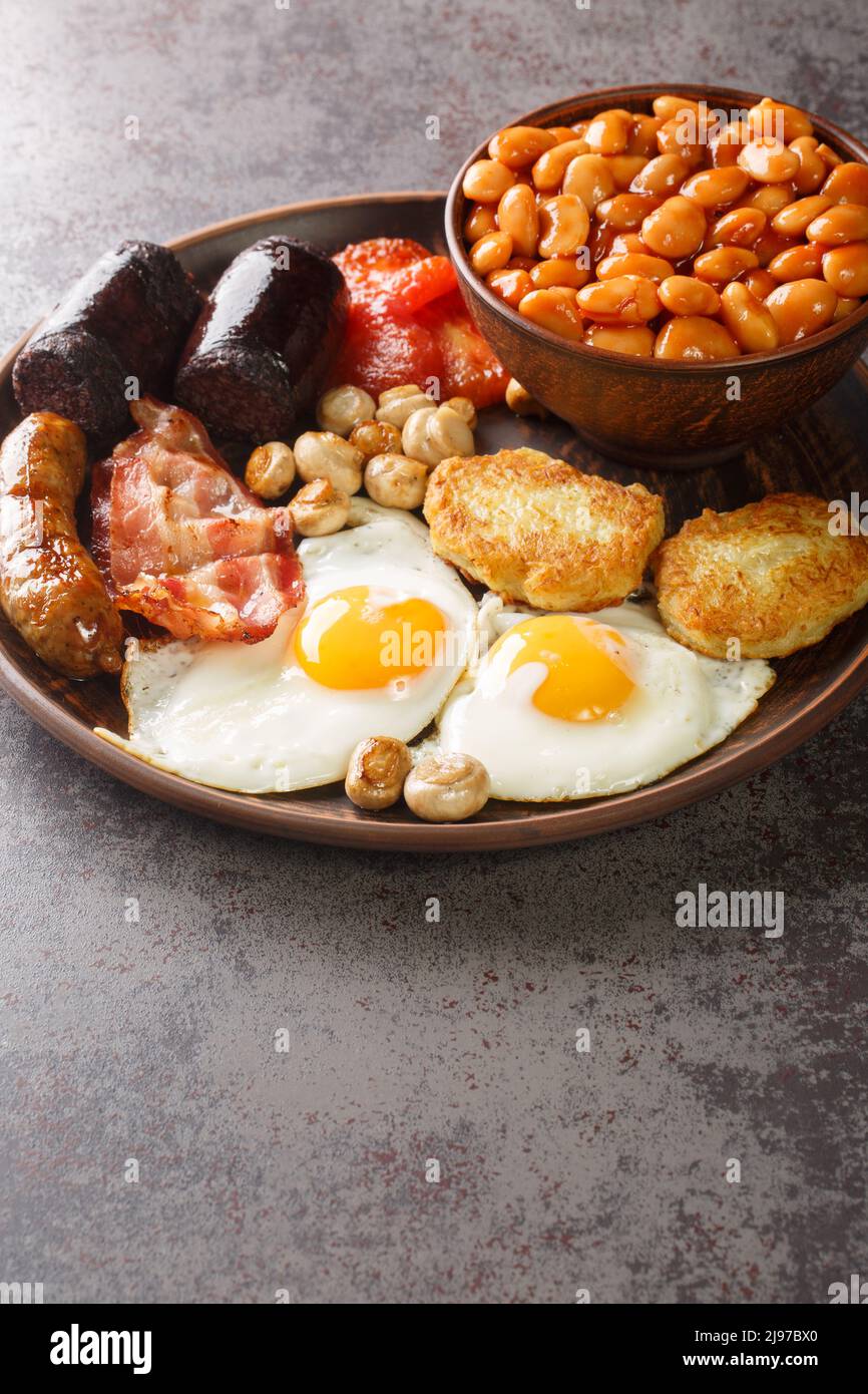 Komplettes englisches Frühstück mit Speck, Wurst, Spiegelei, gebackenen Bohnen, Kartoffelrösti und Pilzen in der Nähe auf dem Teller auf dem Tisch. Vertikal Stockfoto