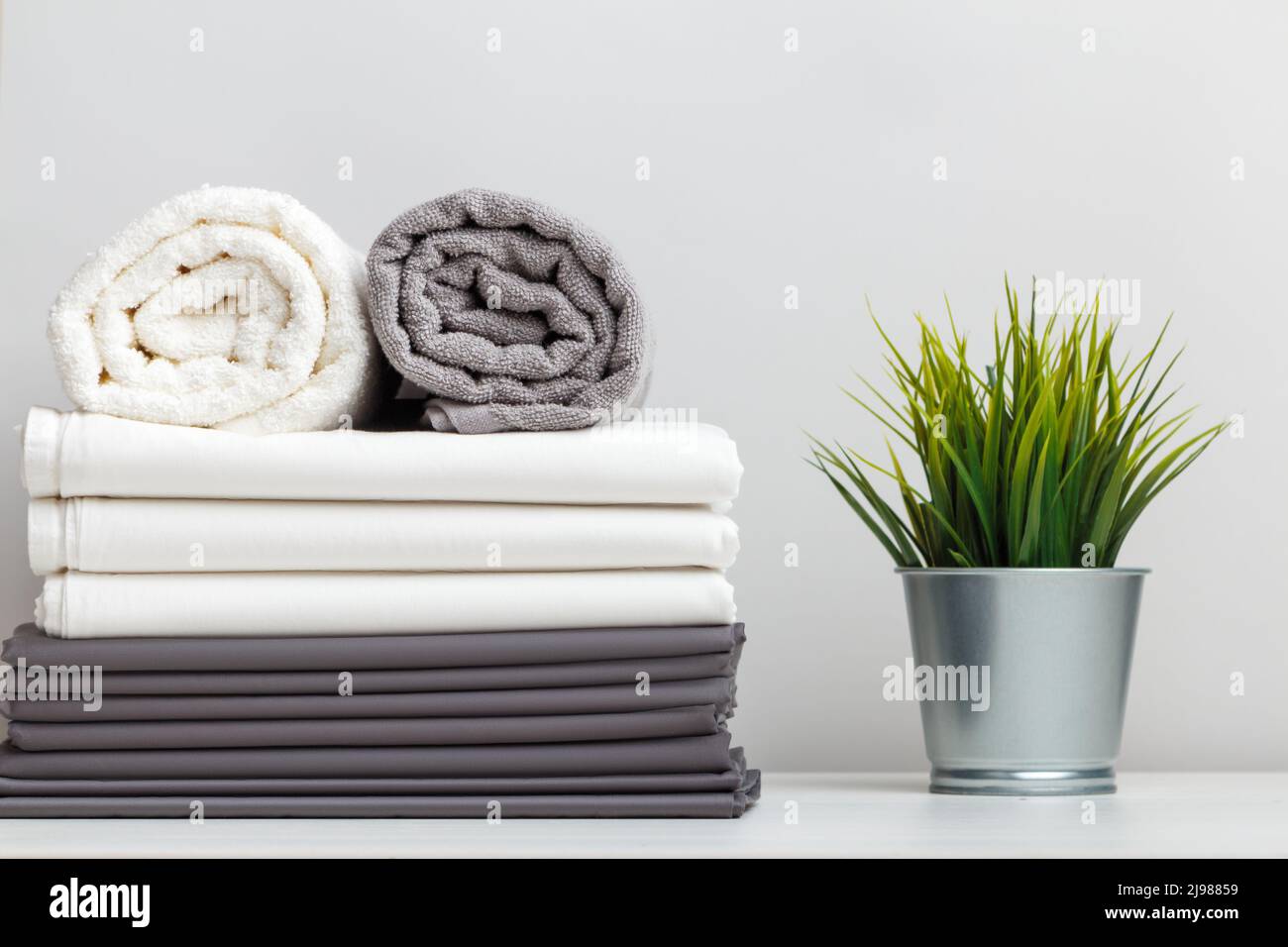 Ein Stapel grauer und weißer Bettwäsche, Bettwäsche und eine gefaltete Rolle Handtücher, eine Pflanze in einem Topf auf einem Tisch. Stockfoto