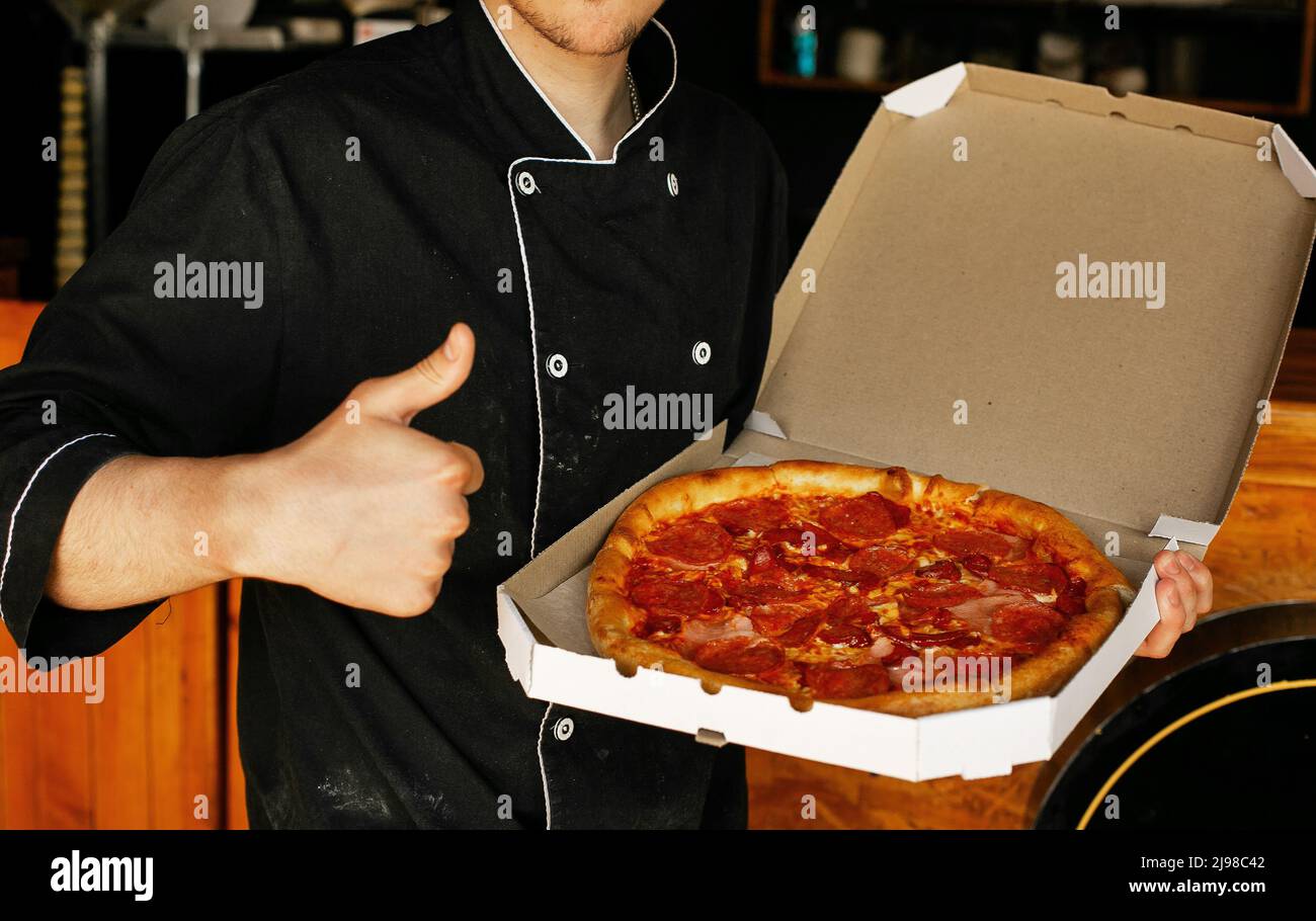 Pizzakoch mit schwarzer Schürze, der in der Pizzeria des Pizzashofs frisch gebackene Pizza in offener Box hält Stockfoto