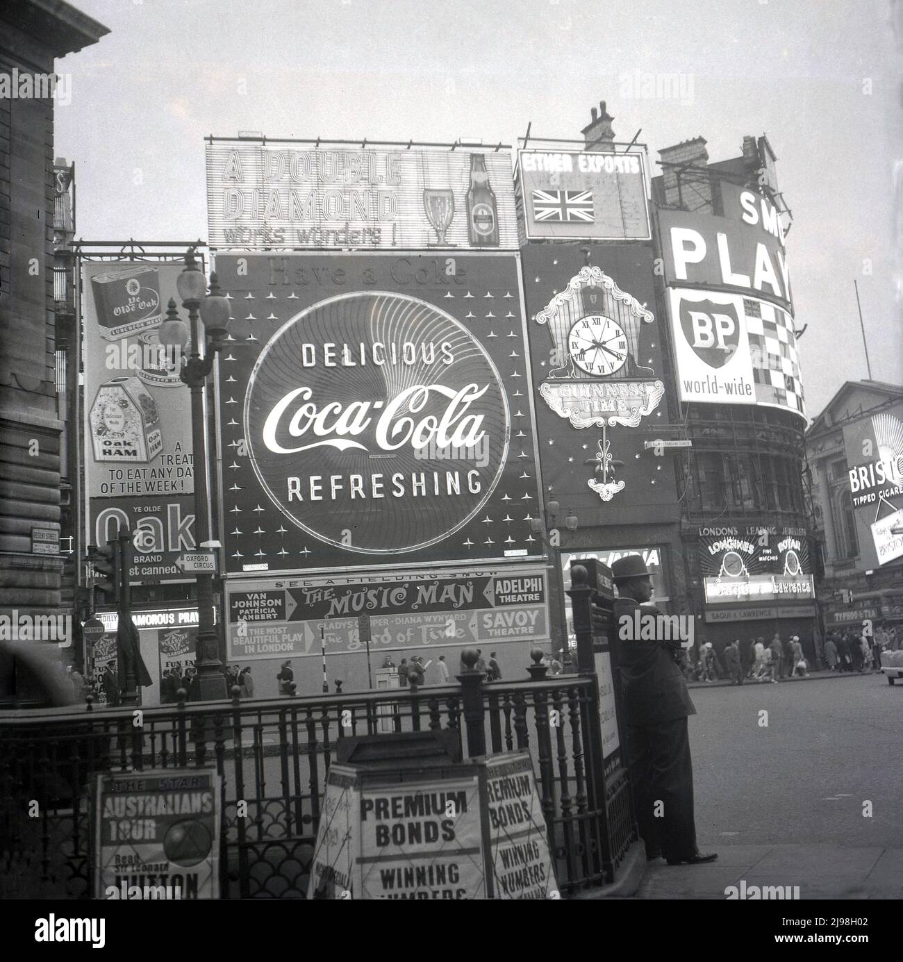 1961, historischer Mann in einem Hut, der am Geländer im Piccadilly Circus, London, England, Großbritannien, steht, mit den berühmten Neon-Werbetafeln, die die Werbetreibenden und Marken des Tages zeigen, darunter Ye Olde Oak Ham, Double Diamond Beer, Players Cigarettes und BP. Das Musical von Meredith Willson, The Music man, ist im Adelphi Theater im West End zu sehen. Stockfoto