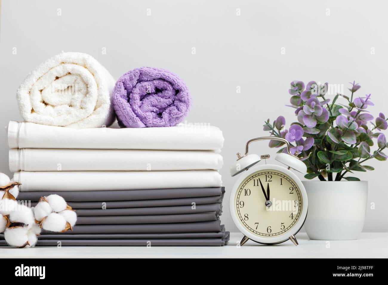 Stapel Bettwäsche, Bettwäsche und Handtücher mit Baumwollzweig und Wecker mit Topfpflanzen auf dem Tisch Stockfoto