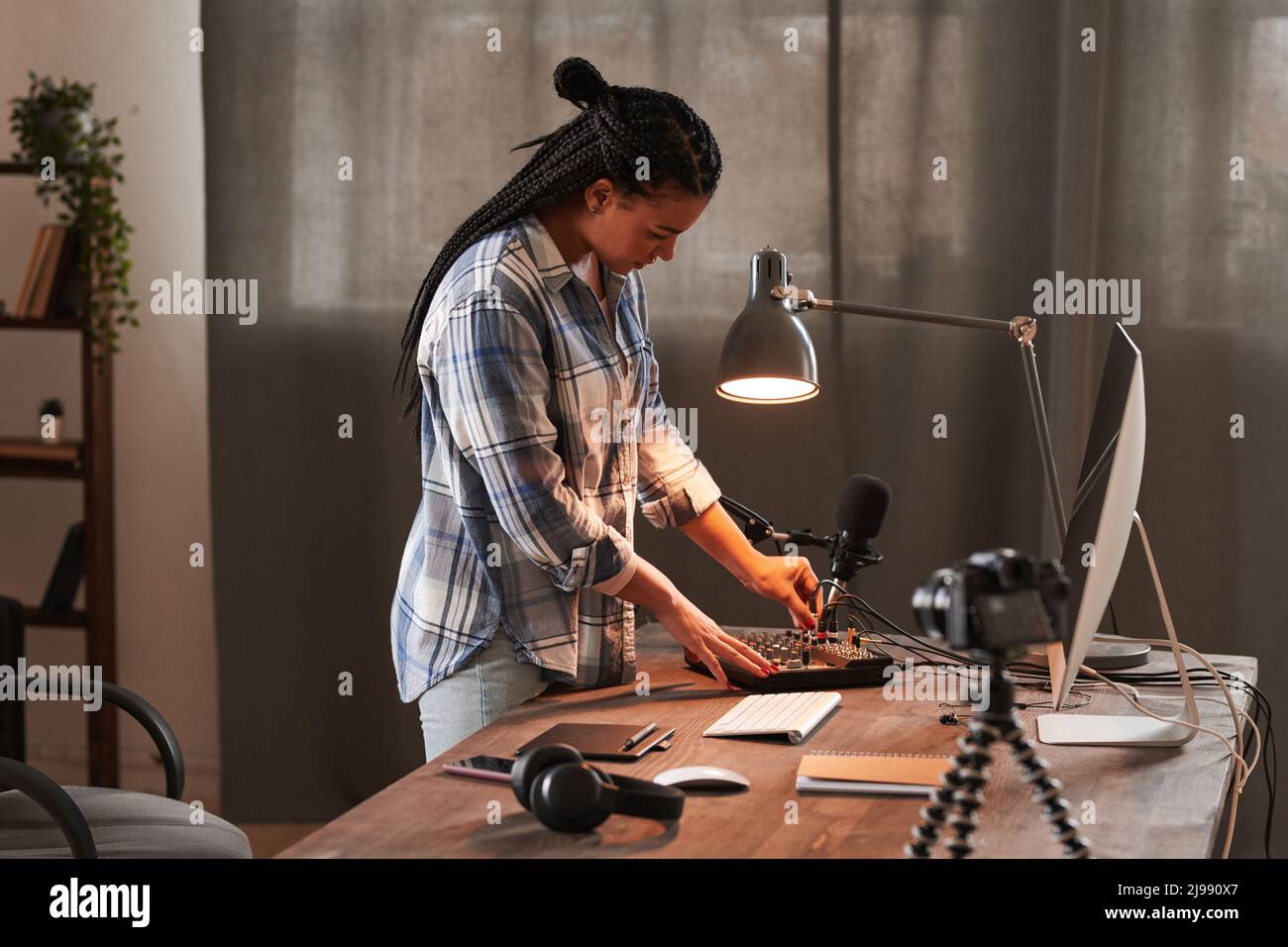 Stilvolle junge Frau, die am Tisch im Loft-Home-Office-Raum steht und das Mikrofon anmacht, um sich auf das Vlogging vorzubereiten Stockfoto