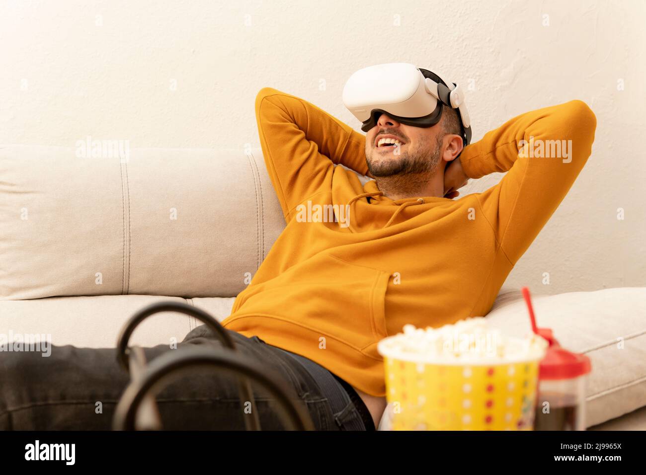 Mann, der auf der Couch sitzt und einen Film in seiner Virtual-Reality-Brille mit Popcorn und Softdrink anschaut Stockfoto