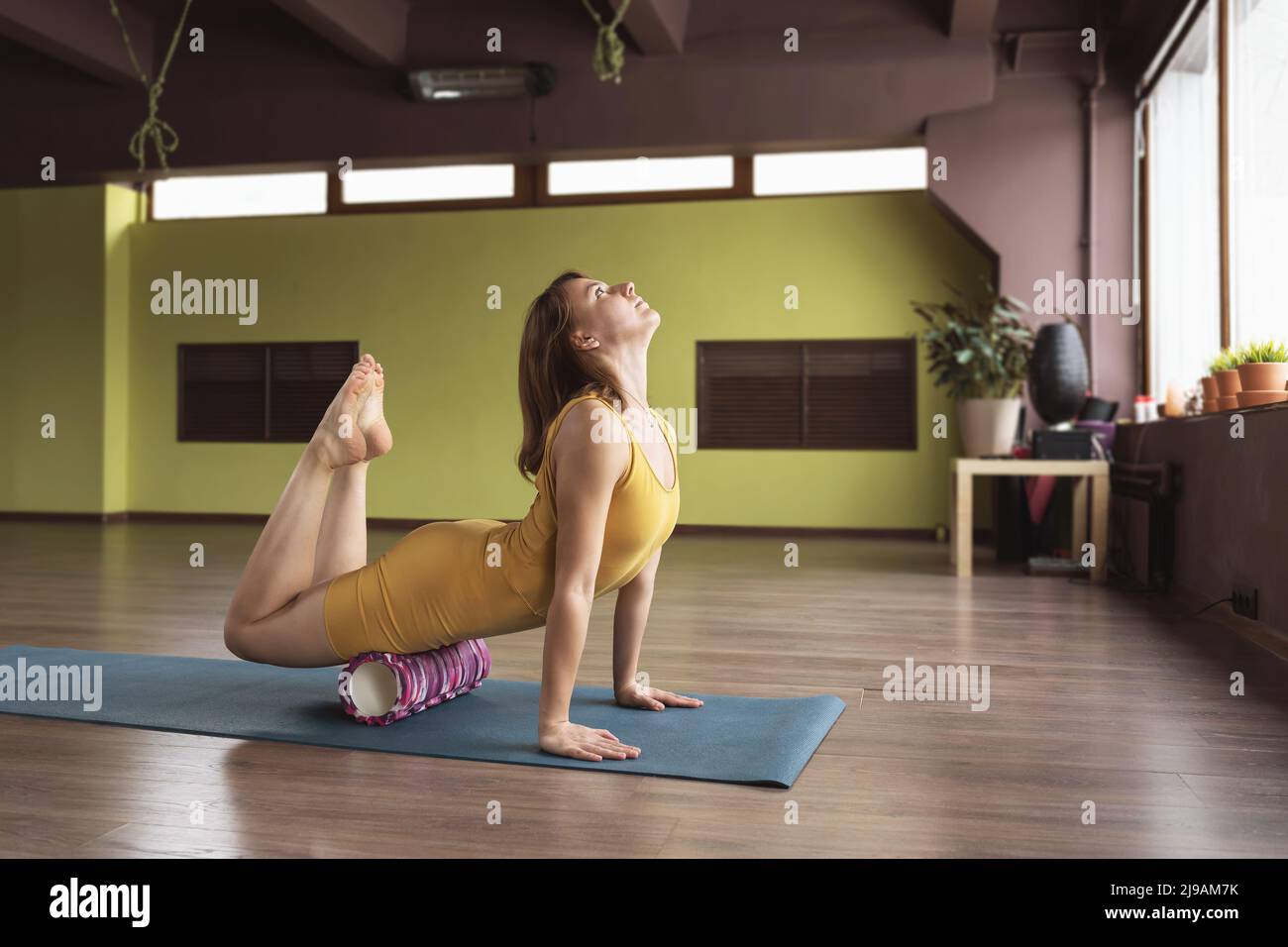Eine Frau in Sportbekleidung führt mit einem Roller eine myofasziale Massage der Beinmuskulatur durch, trainiert in einem Raum mit einer Rückbiegung auf einer Matte Stockfoto