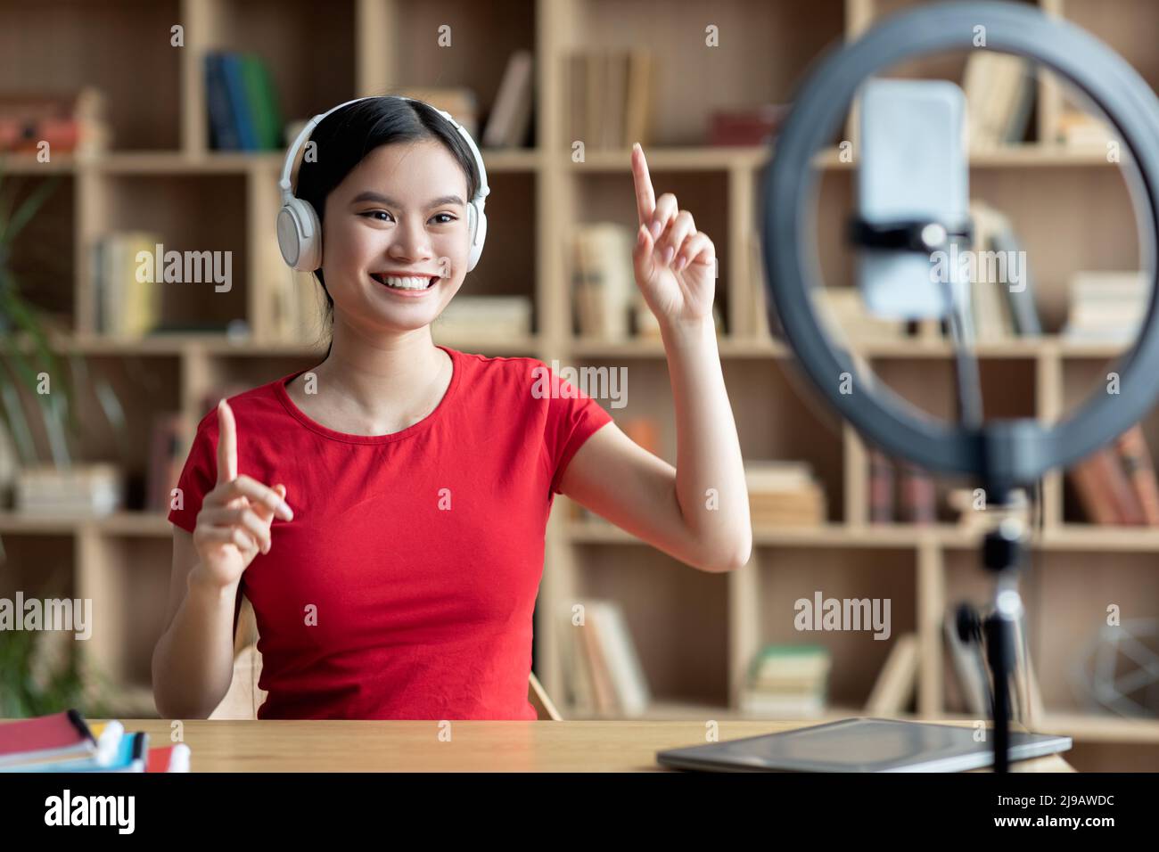 Glückliche junge asiatische Frau in kabellosen Kopfhörern gestikuliert mit den Händen und Blick auf Smartphone-Webcam Stockfoto